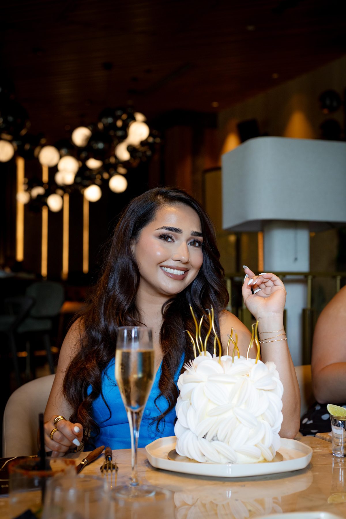 Birthday celebration photograph of a woman lighting the candles on a white ruffled cake at a restaurant table, champagne glass beside her