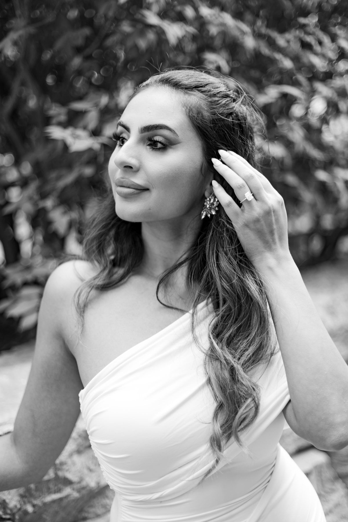 Black and white outdoor portrait of the bride-to-be in a draped white gown, tucking her hair back against a soft garden background