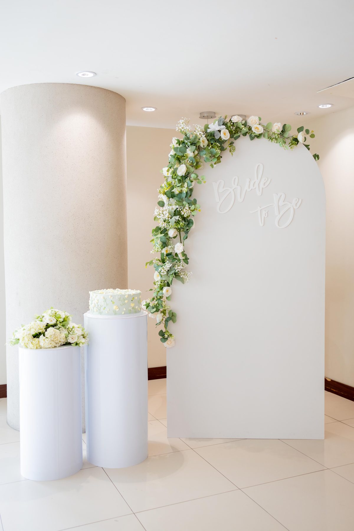 Bridal shower decor detail: a white arch backdrop trailing eucalyptus and white roses beside a two-tier cake on a white plinth