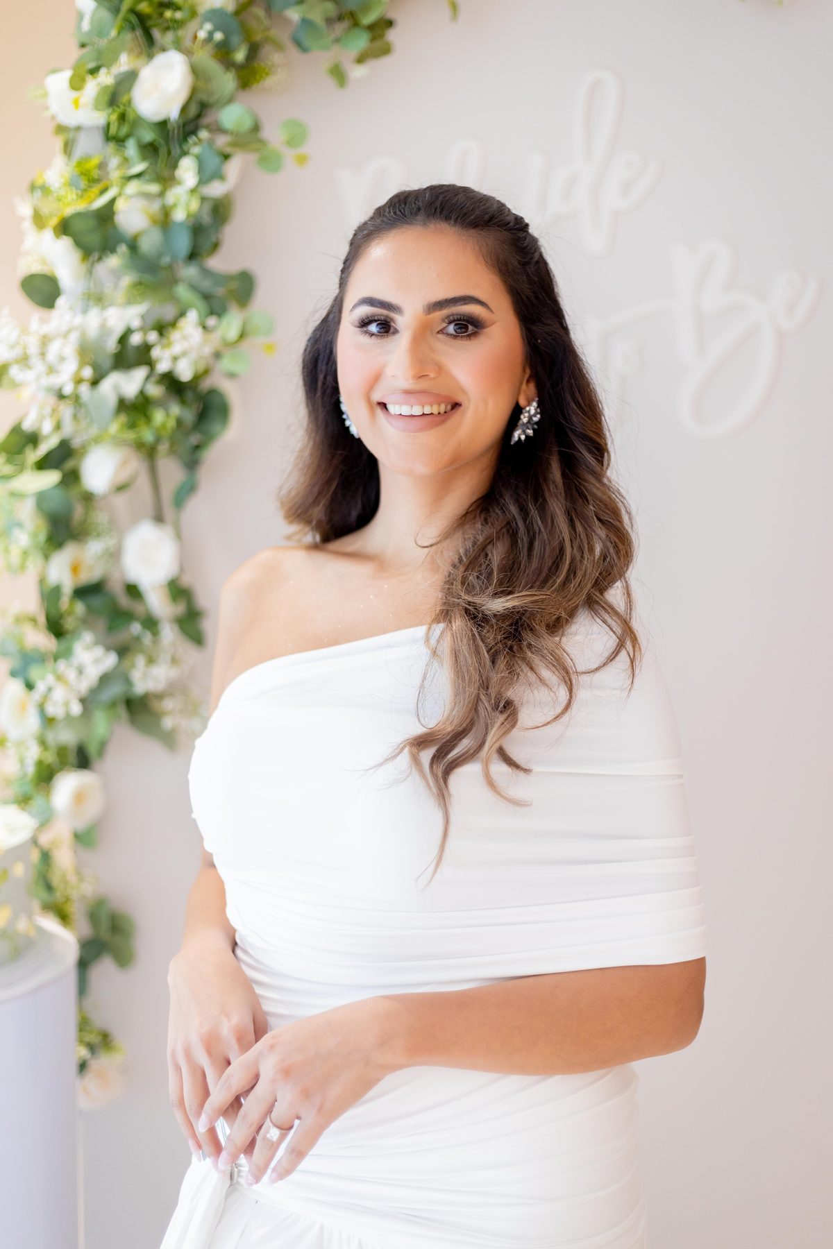 Portrait of the bride-to-be in a white one-shoulder gown standing in front of the floral bridal shower backdrop