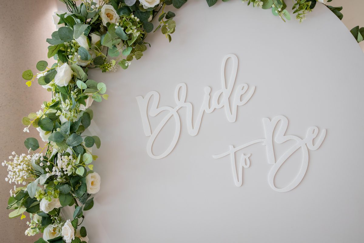 Bridal shower backdrop with a hand-lettered Bride To Be sign framed by a greenery and white rose arch
