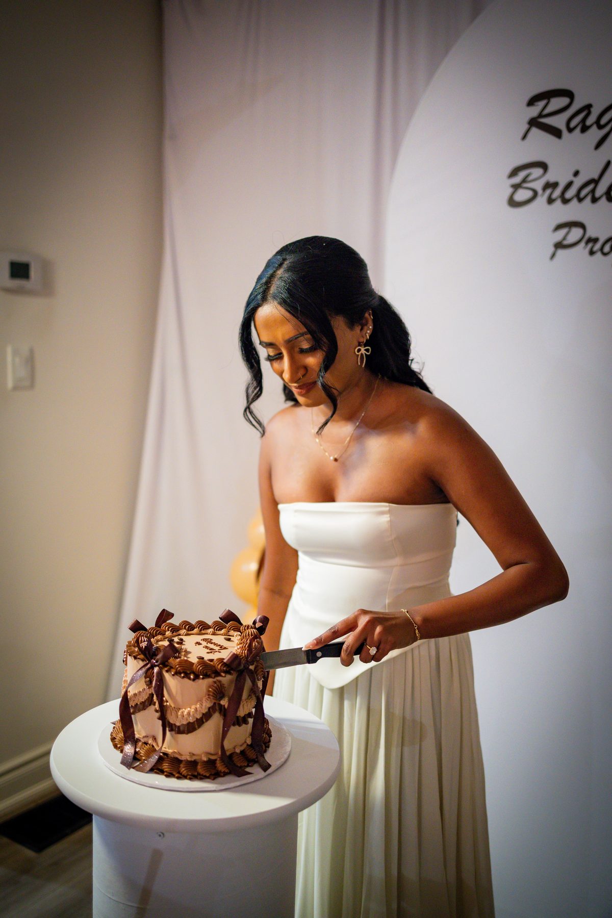 Bride-to-be in a white gown cutting a chocolate drip cake at her bridesmaid proposal party