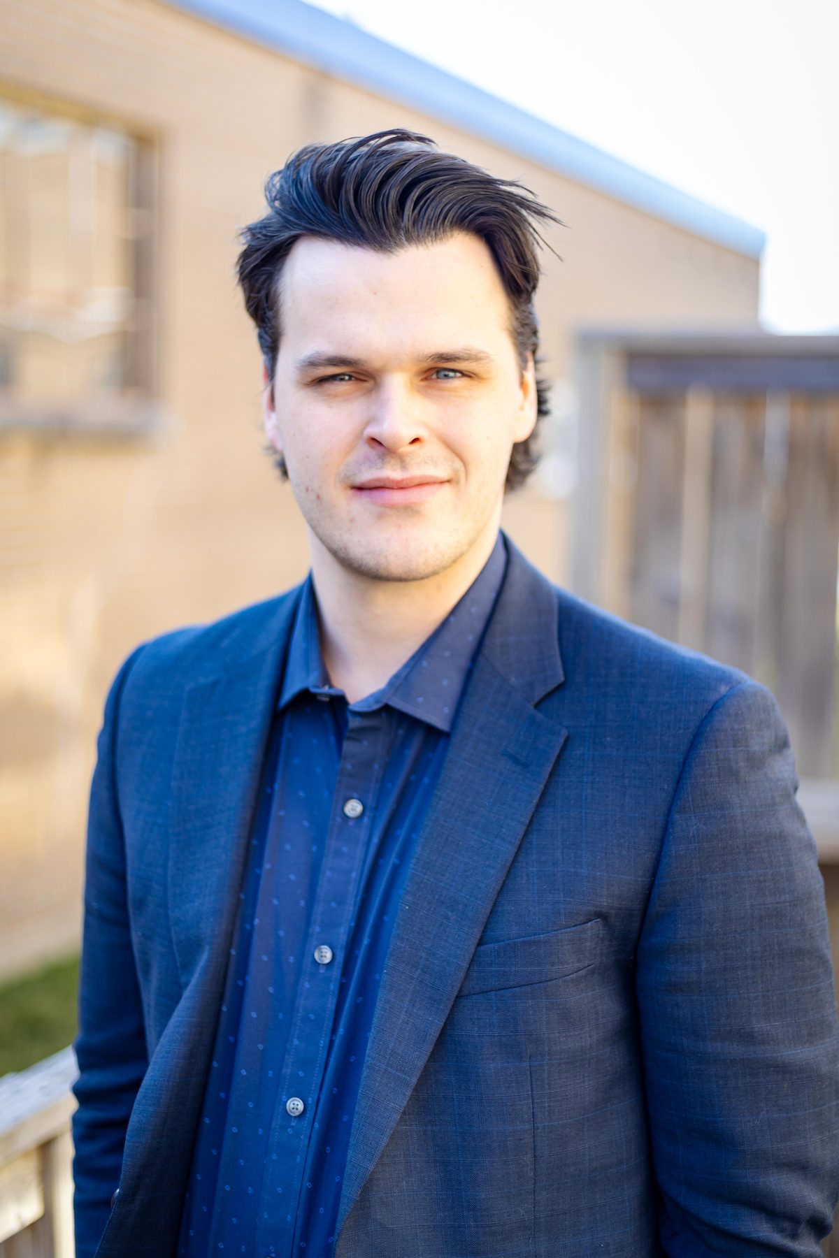Business headshot of a man in a blue blazer over a navy shirt, photographed outdoors against a soft architectural background