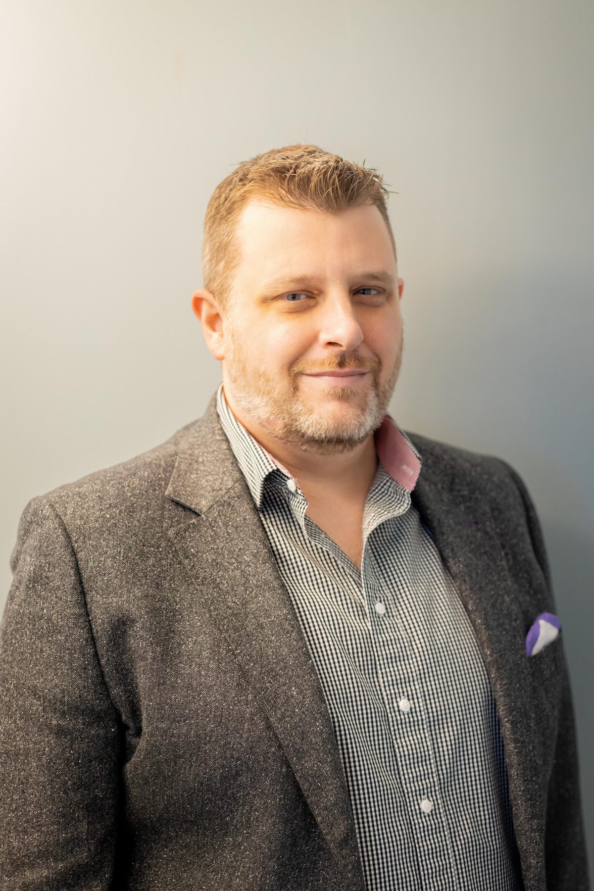 Corporate headshot of a man in a tweed blazer and checked shirt with a pocket square, shot against a soft grey backdrop