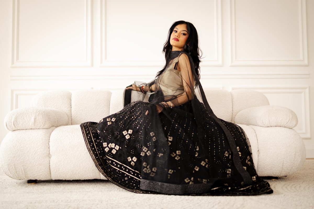 Editorial portrait of a woman in a black embellished gown seated on a cream sofa in a bright panelled studio