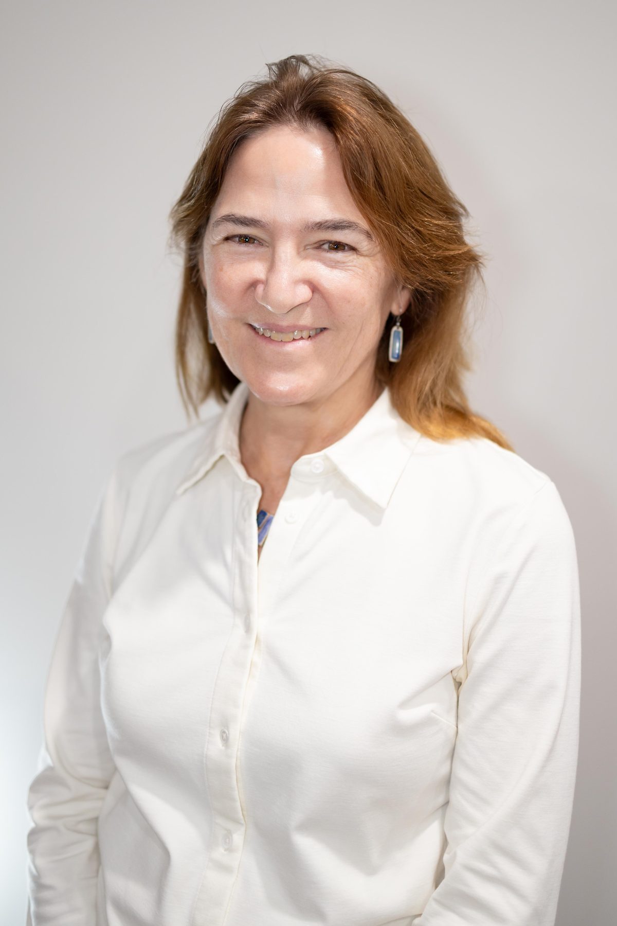 Executive headshot of a smiling woman in a crisp white blouse, lit by warm window light against a neutral wall