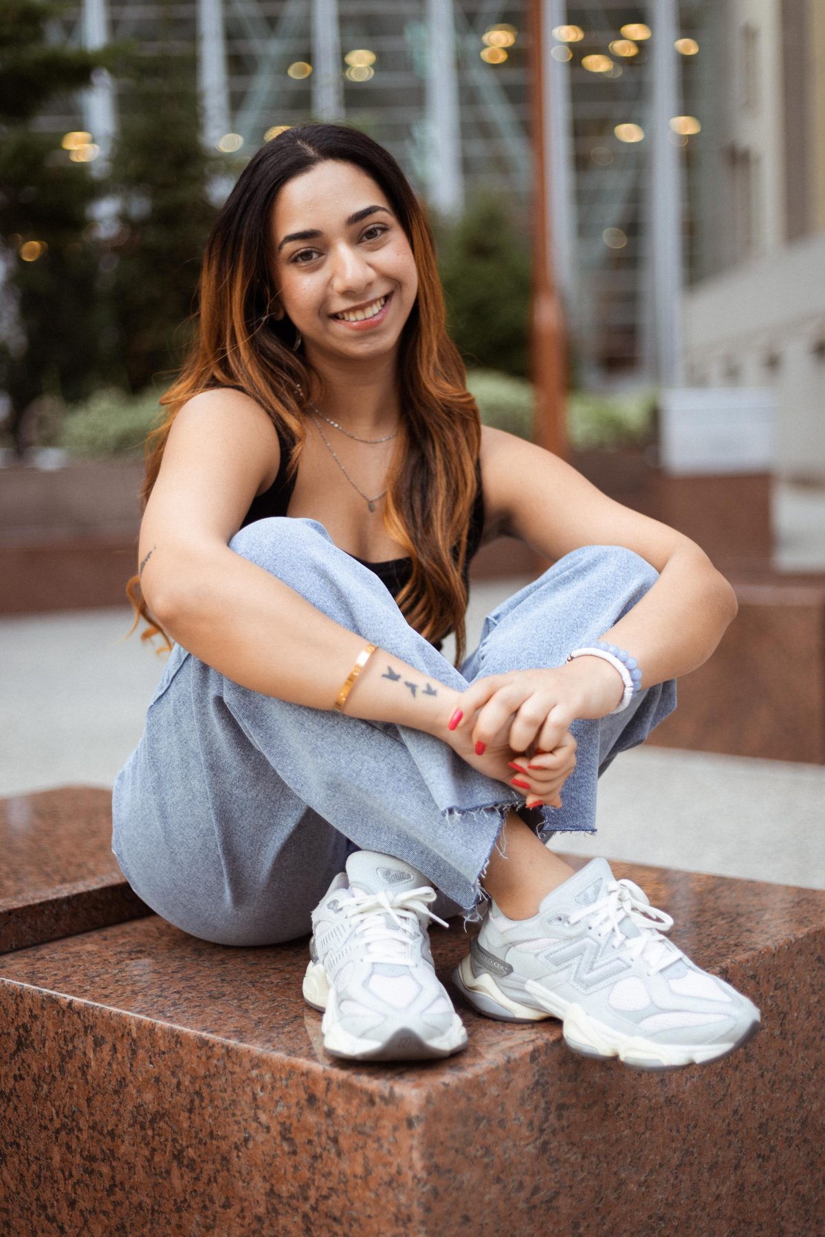 Relaxed lifestyle portrait of a woman seated cross-legged on a granite ledge in denim and white sneakers, smiling at the camera