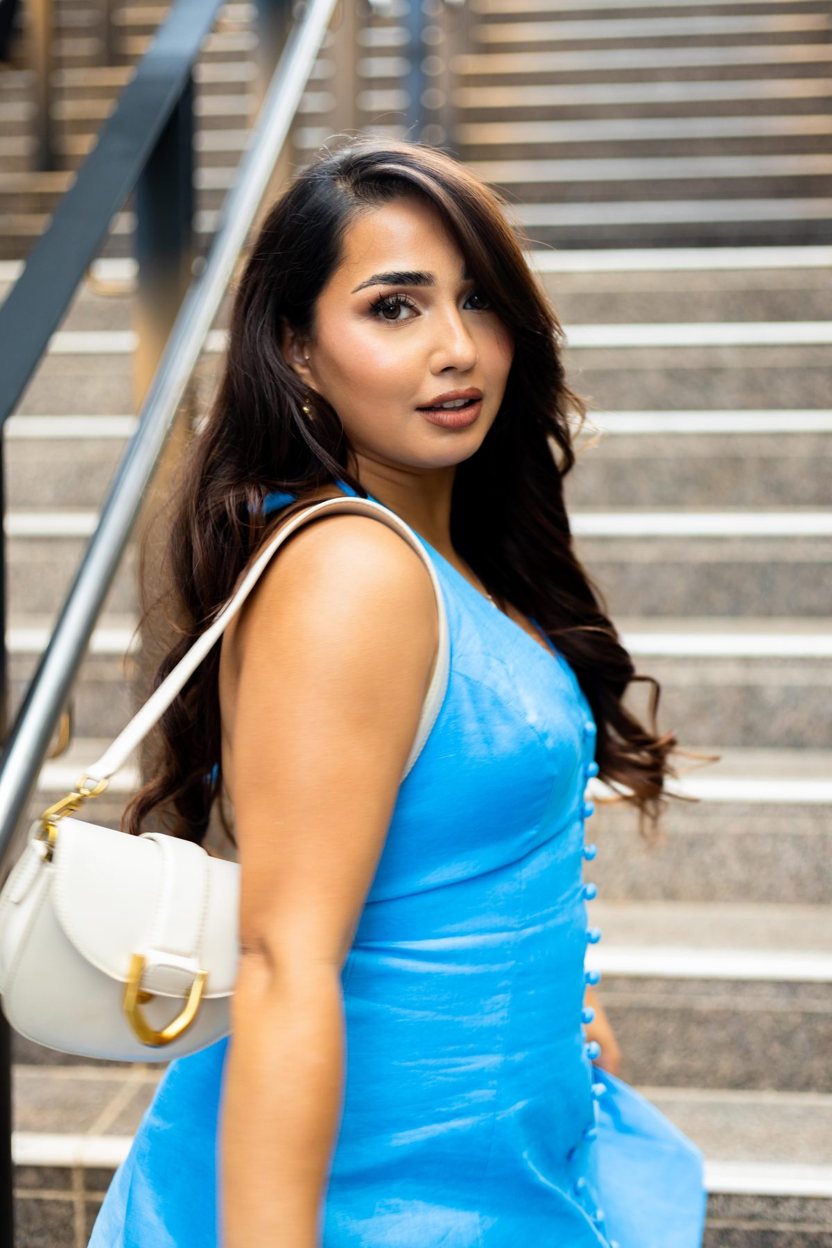 Outdoor portrait of a woman in a blue satin dress with a white shoulder bag, looking back over her shoulder on a wide stone staircase