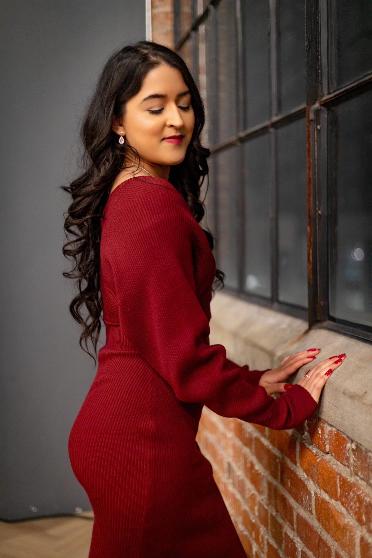 Portrait of a woman in a red knit dress turning toward a tall industrial window, hand resting on the brick sill