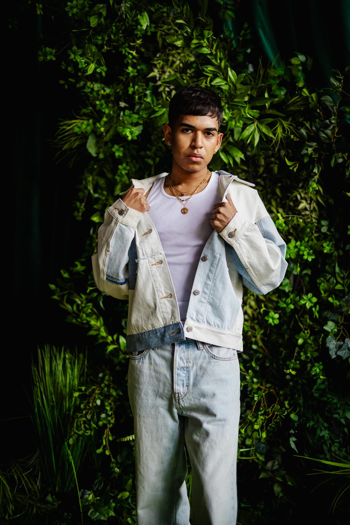Portrait of a young man in a denim jacket and layered chains standing against a dark green foliage backdrop