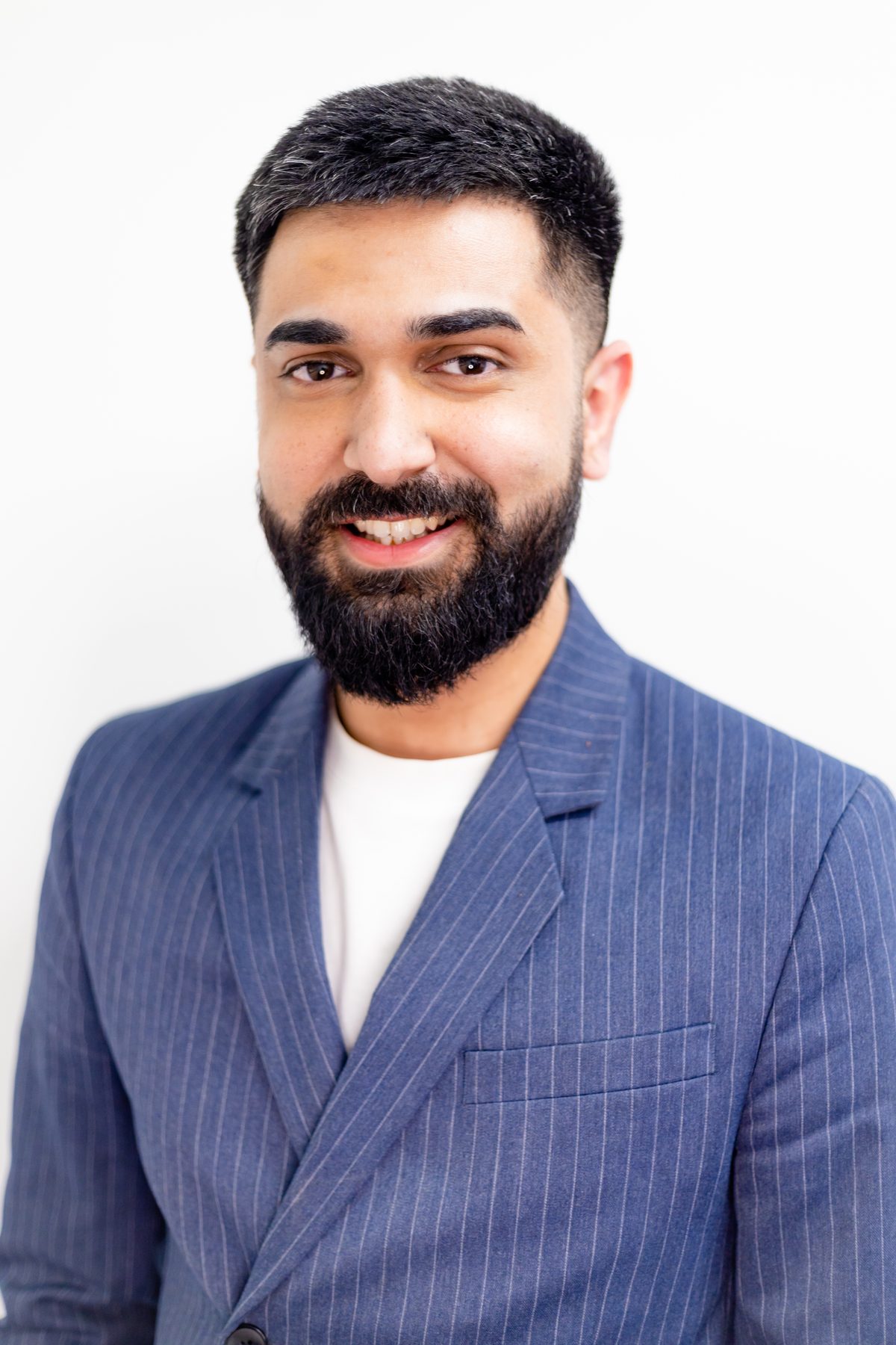 Professional headshot of a bearded man in a blue pinstripe blazer over a white tee, shot on a white studio backdrop