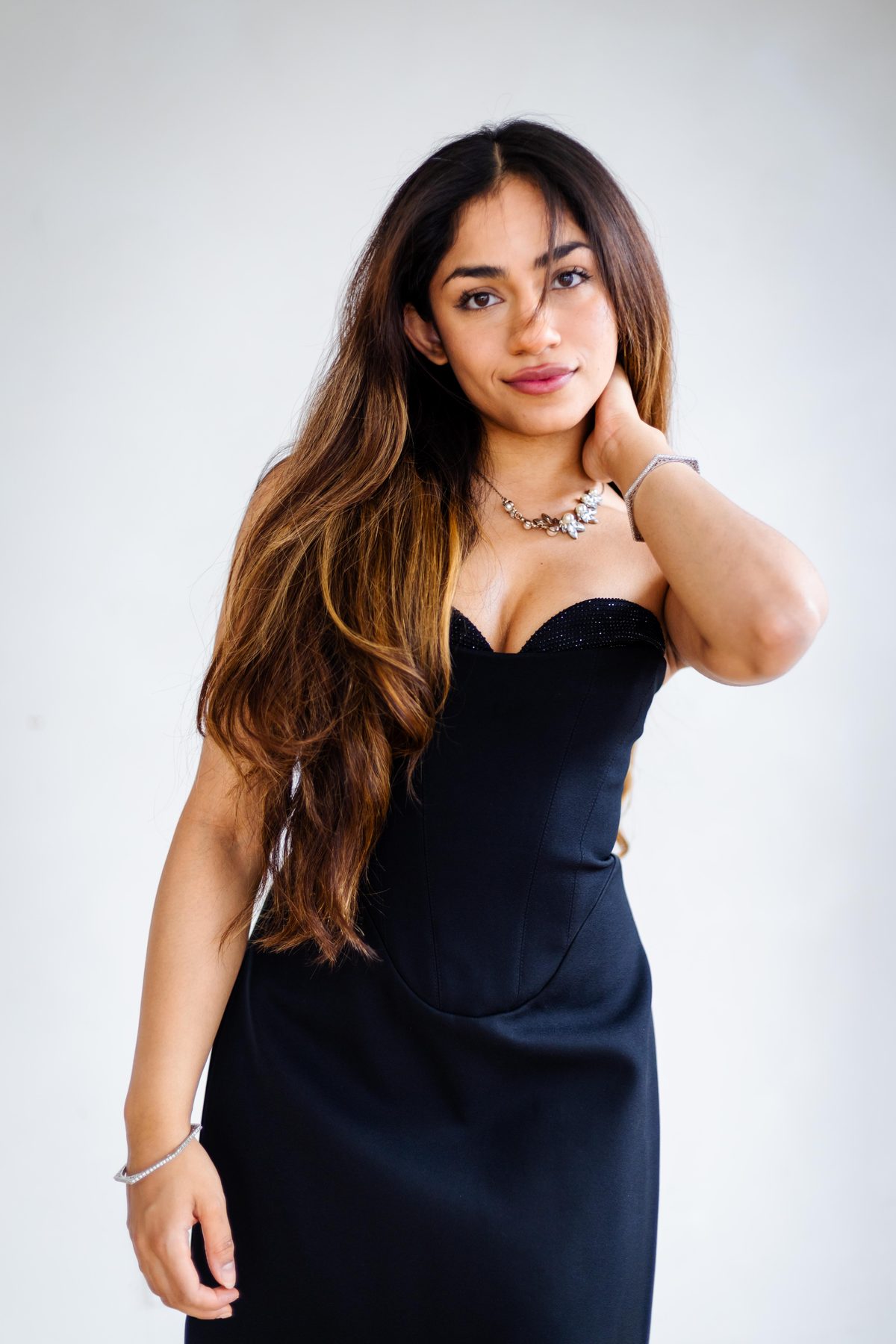 Studio portrait of a woman in a black corset evening gown against a clean white backdrop, hand resting at her neck