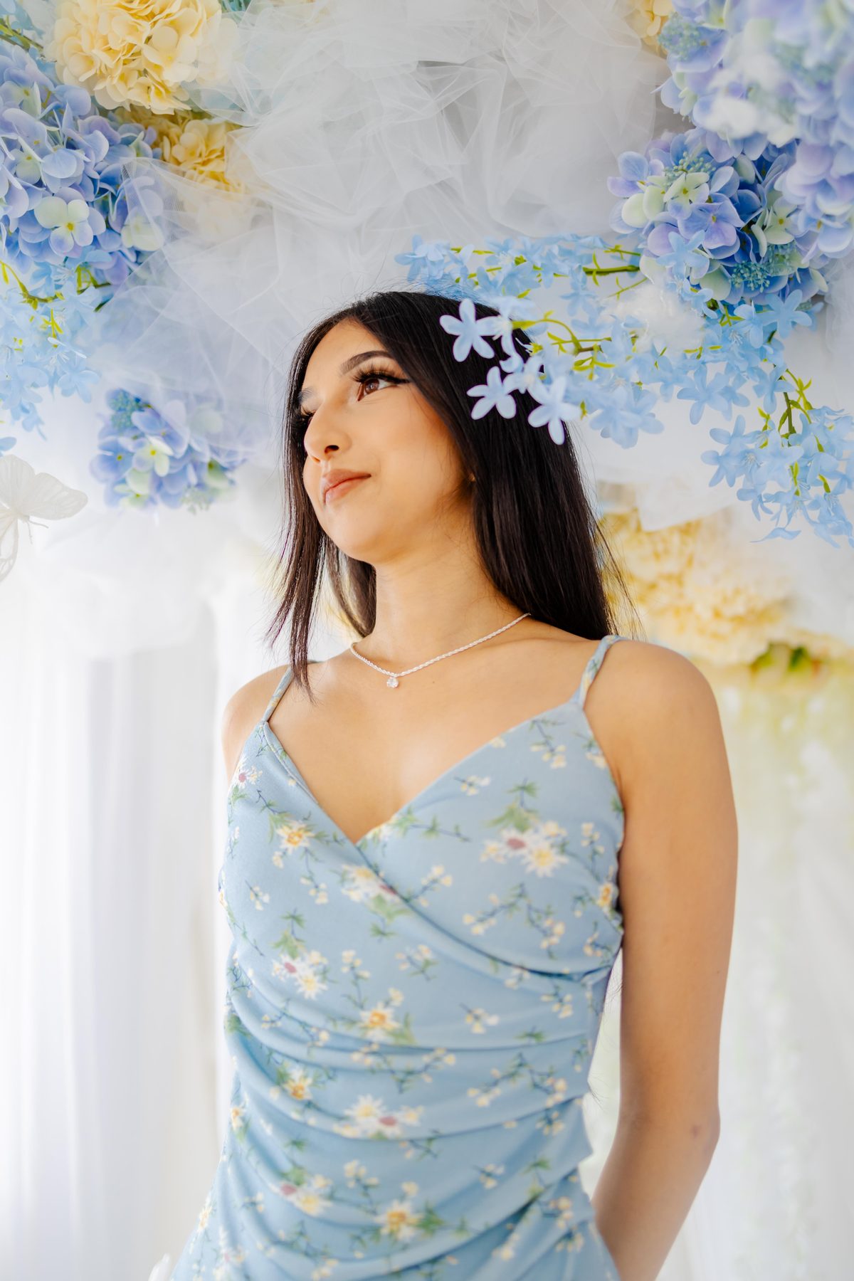Styled portrait of a young woman in a blue floral dress looking upward beneath a pale blue hydrangea arch
