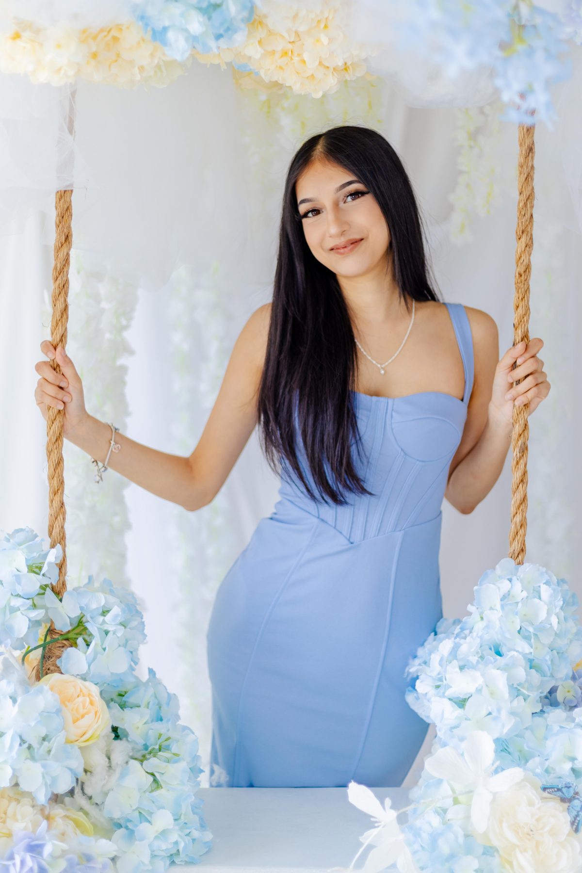 Styled portrait of a young woman in a blue dress seated on a rope swing dressed with pale blue hydrangeas